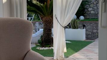 A beige armchair in a patio area with a view of a garden, a palm tree, and a stone wall.