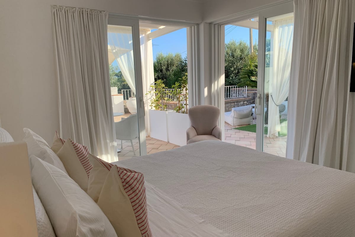 A bedroom with a large bed, white curtains, and a view of an outdoor patio with a chair and plants.