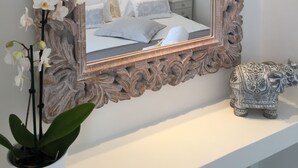 Un dormitorio con un espejo grande y ornamentado, una cama con ropa de cama blanca, una mesita de noche pequeña y una figurita decorativa de elefante.