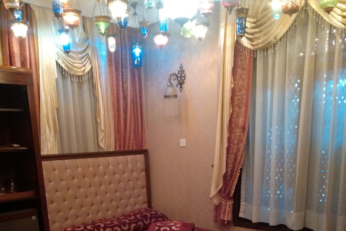 Quarto com cabeceira estofada, cama grande com roupa de cama estampada, mesa de cabeceira com velas e janela com cortinas.