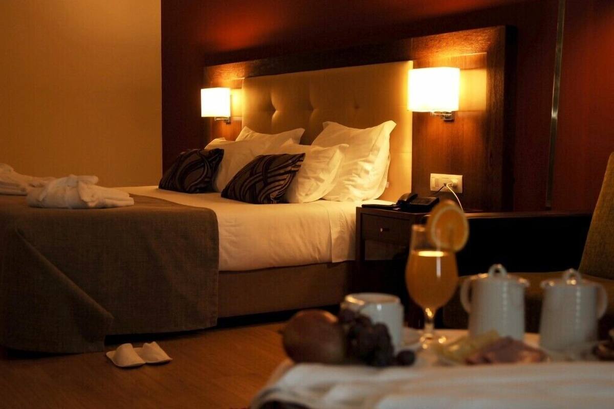 A hotel room with a bed, bedside lamps, a nightstand, a mirror, and a breakfast setup with a glass of orange juice and two mugs.