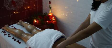 Sesión de masoterapia en un spa, con una persona acostada en la camilla, una pared decorativa y una planta con velas.