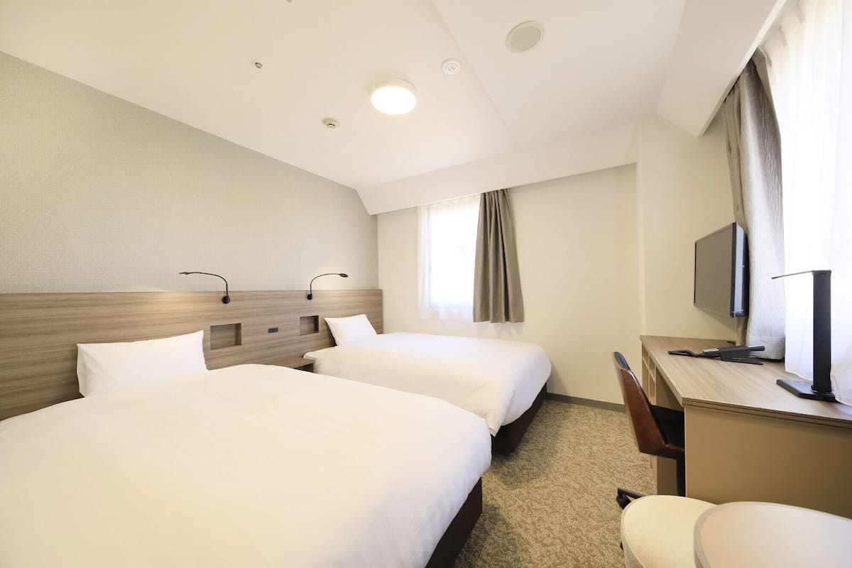 A hotel room with two beds, a desk, a chair, a television, and a window with curtains.
