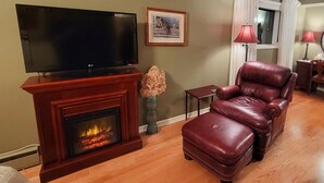 A living room with a flat-screen TV on a wooden stand, a fireplace with a lit fire, a leather armchair, a small table, and a framed picture on the wall. - Forest Hill Bed & Breakfast (Kitchener)