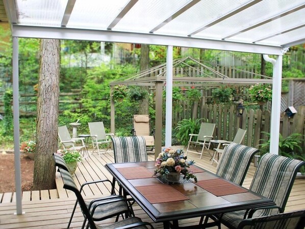 Une terrasse couverte avec une table et des chaises, entourée d’une terrasse en bois et d’un jardin.