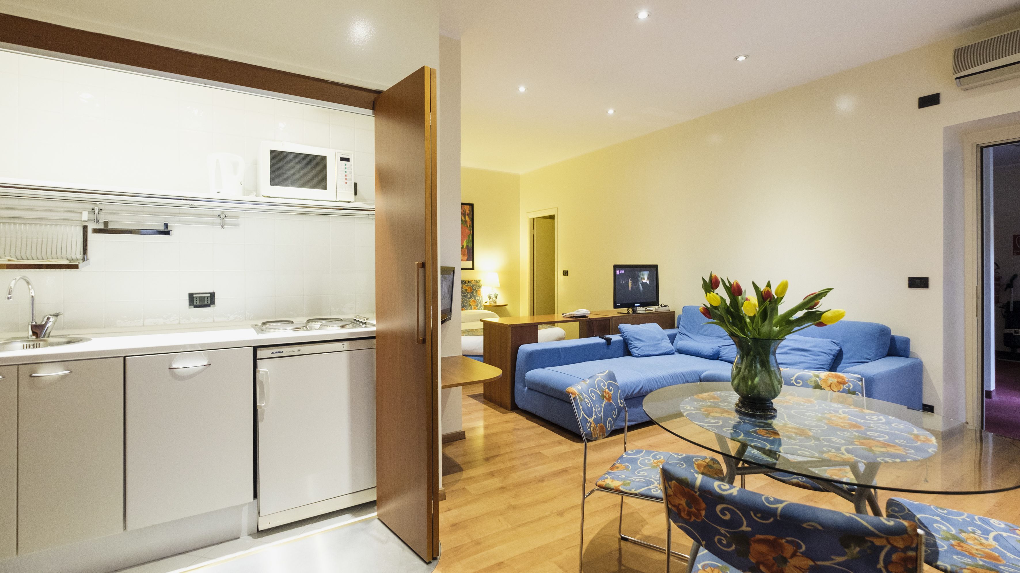 Cocina moderna con microondas, estufa y lavavajillas. Hay una sala de estar con un sofá y una mesa de centro de vidrio. Un jarrón con flores está sobre la mesa.
