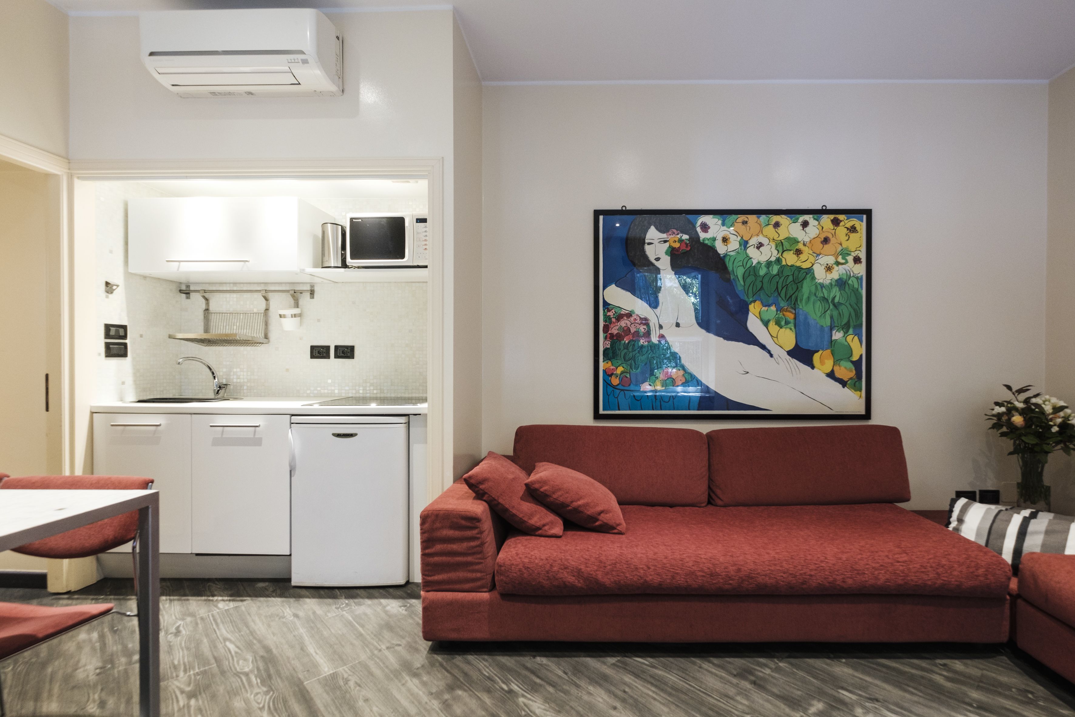 A modern kitchen with white cabinets, a microwave, and a dishwasher. There is a red sofa and a framed painting on the wall.