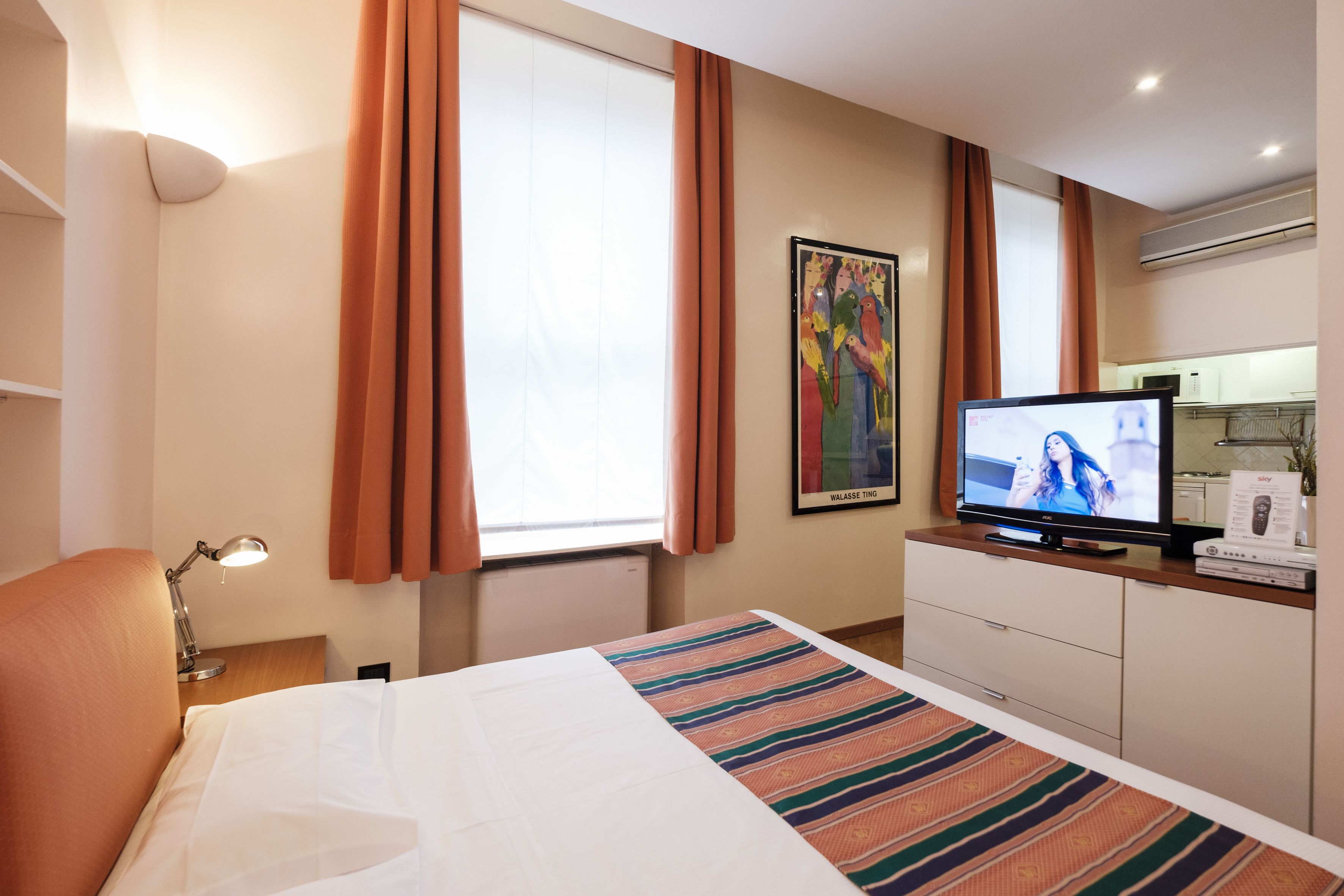 Habitación de hotel con cama, televisión, escritorio con lámpara, un cuadro en la pared y ventana con cortinas.