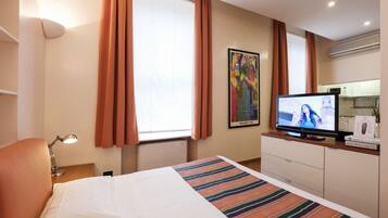 Habitación de hotel con cama, televisión, escritorio con lámpara, un cuadro en la pared y ventana con cortinas.