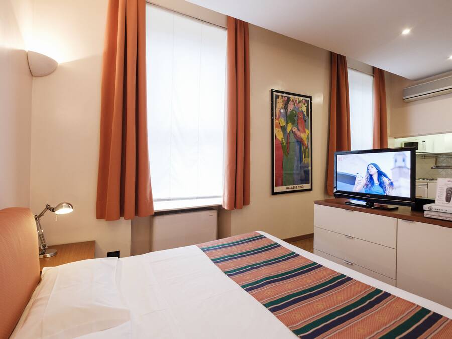 Habitación de hotel con cama, televisión, escritorio con lámpara, un cuadro en la pared y ventana con cortinas.