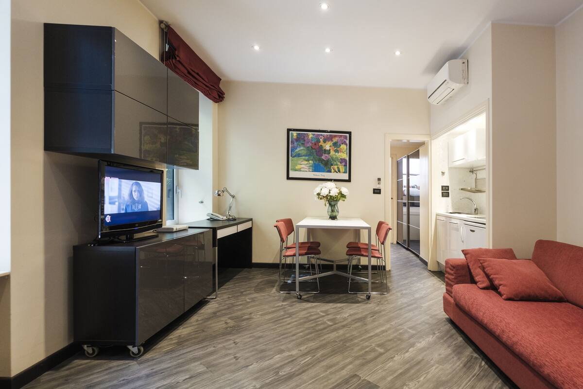 Un soggiorno moderno con un divano rosso, una TV su un mobile, un'area pranzo con tavolo e sedie e una cucina sullo sfondo.