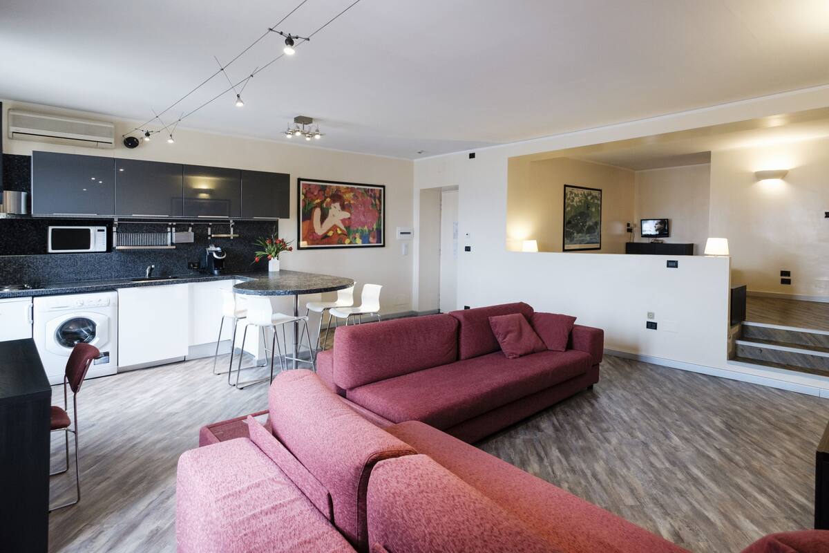 Un soggiorno moderno con un divano rosso, una zona pranzo con un tavolo e sedie, una cucina con mobili scuri e una scala che conduce a un altro piano.