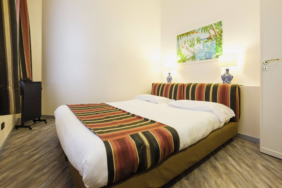 Una camera d'albergo con un letto, un comodino con due lampade, un quadro appeso al muro e una porta.