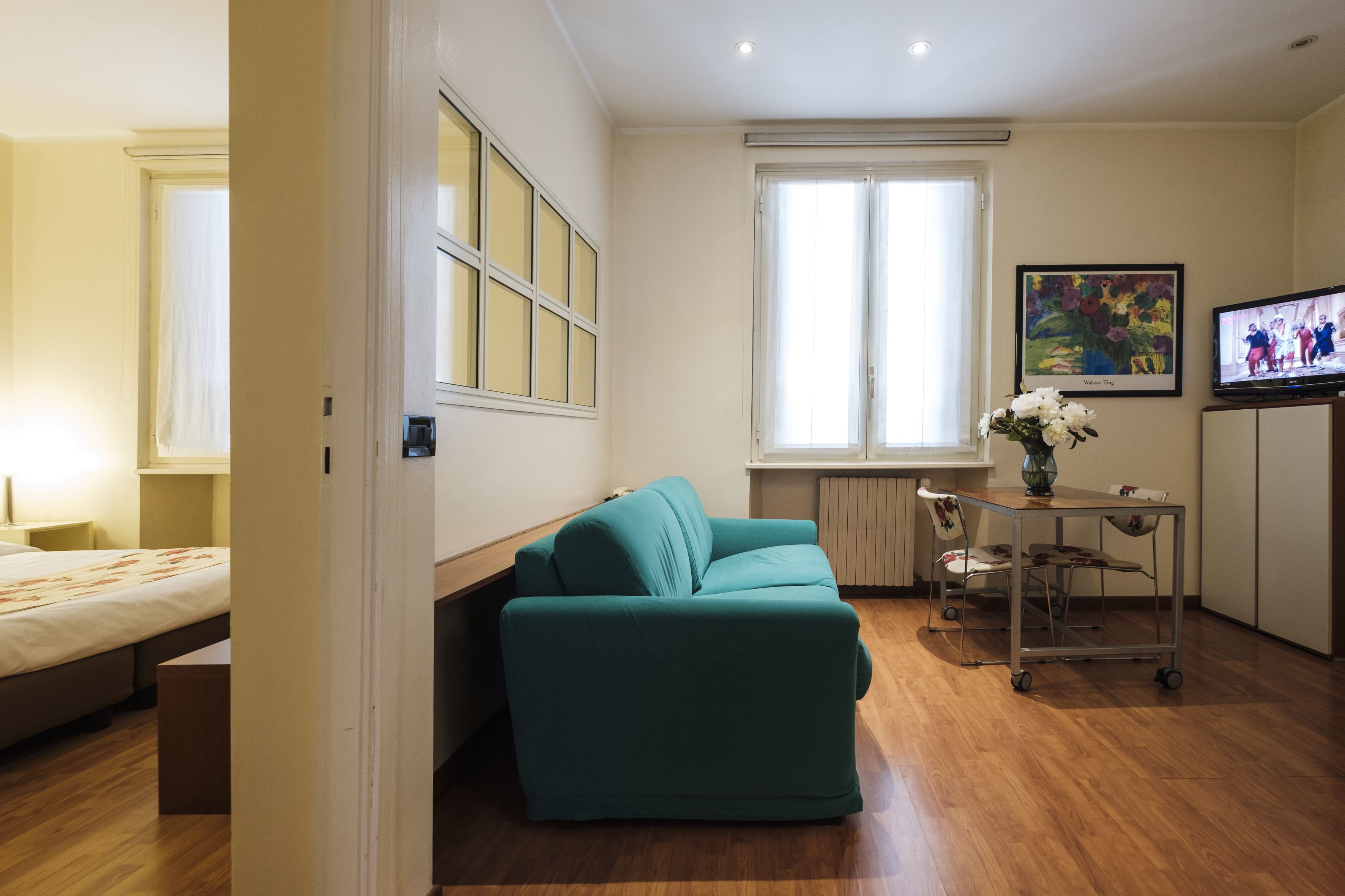 Quarto compacto com sofá verde-água, uma mesinha com um vaso, televisão e cama.