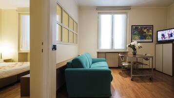 Quarto compacto com sofá verde-água, uma mesinha com um vaso, televisão e cama.