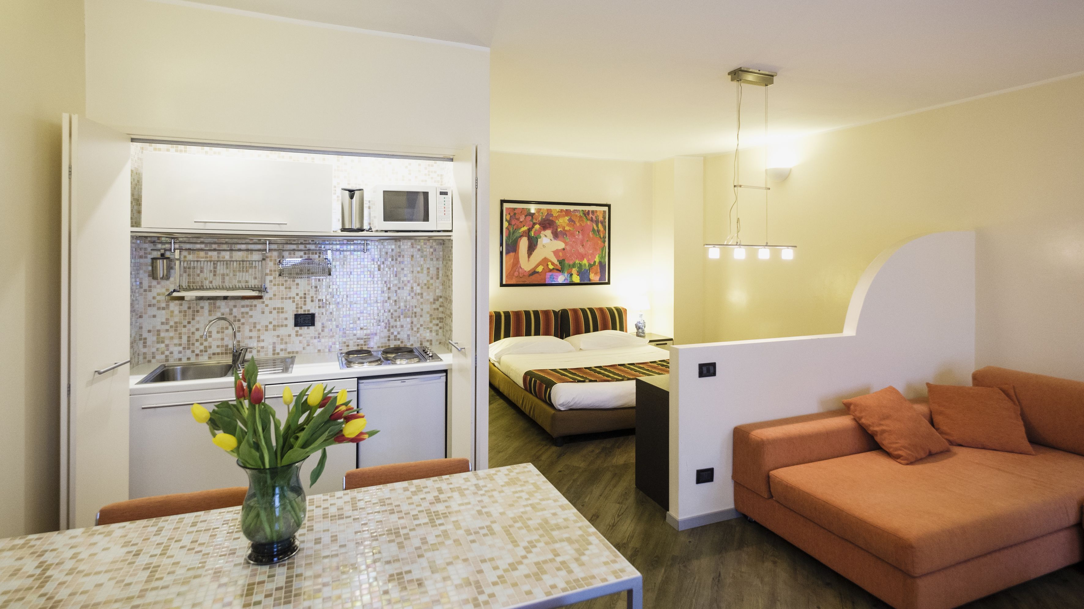 Un espacio habitable compacto con kitchenette, una zona de comedor con un jarrón de tulipanes y un dormitorio con cama y una pintura.