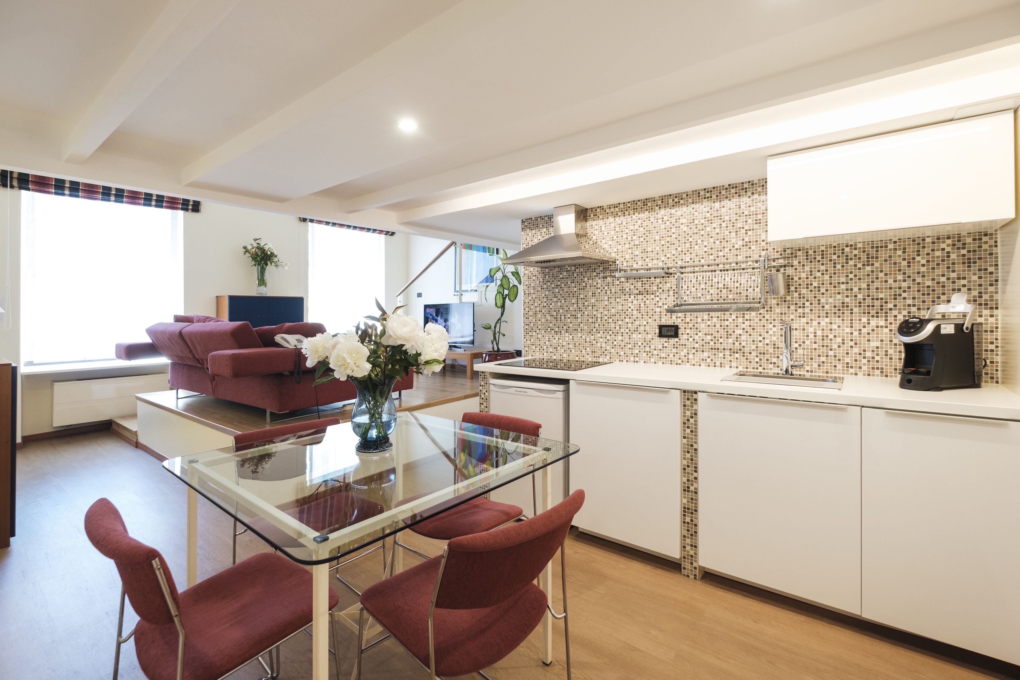 A modern kitchen with a glass dining table, red chairs, and a tiled backsplash.