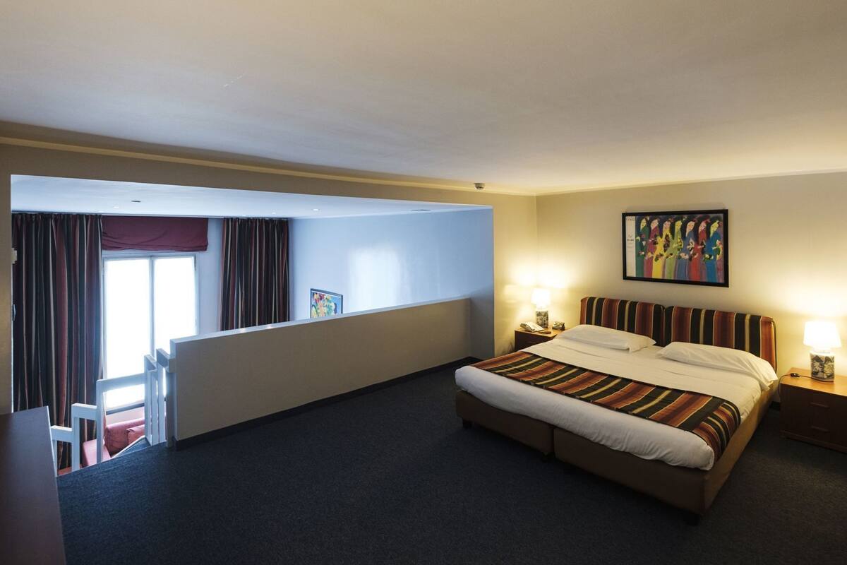 Una camera d'albergo con un letto, abat-jour, un quadro appeso e una finestra con tende.