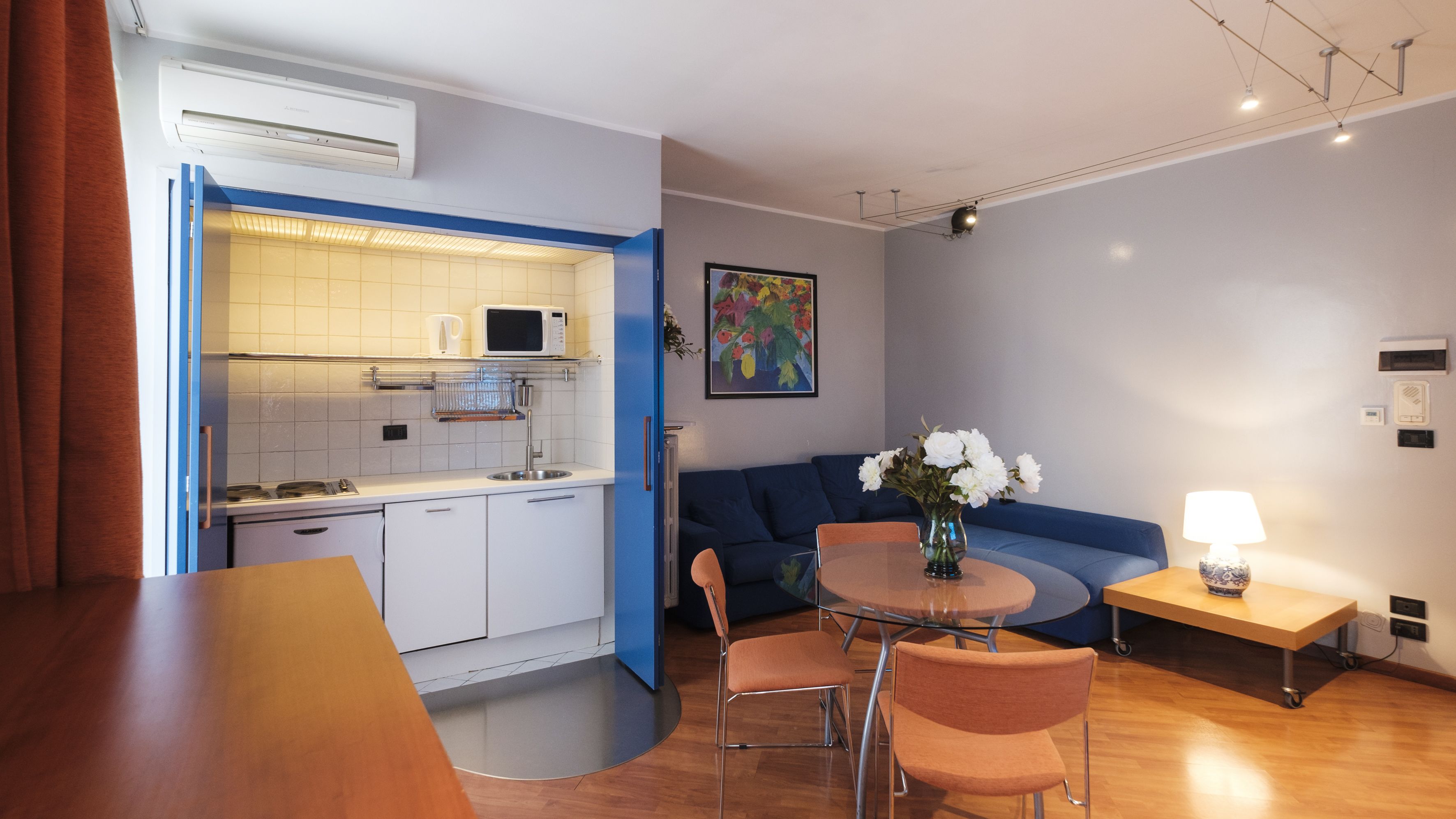 Un espacio habitable compacto con kitchenette, una zona de comedor con mesa y sillas, un sofá y una pequeña mesita de noche con lámpara.