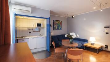 Un espacio habitable compacto con kitchenette, una zona de comedor con mesa y sillas, un sofá y una pequeña mesita de noche con lámpara.