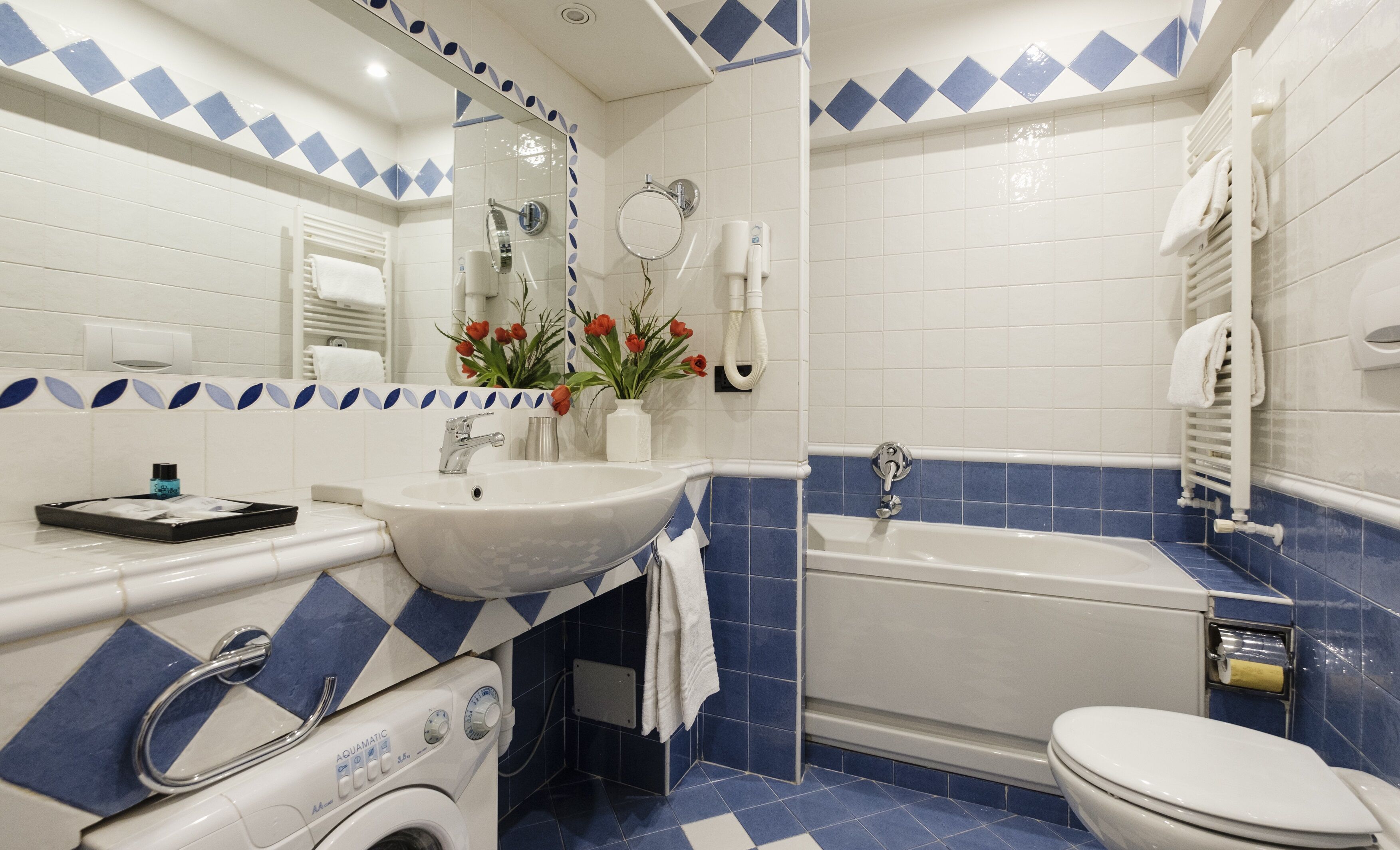 Ein Badezimmer mit einem weißen Waschbecken, einer Badewanne und einer Toilette. Über dem Waschbecken befindet sich ein Spiegel und an der Wand ein Handtuchhalter.