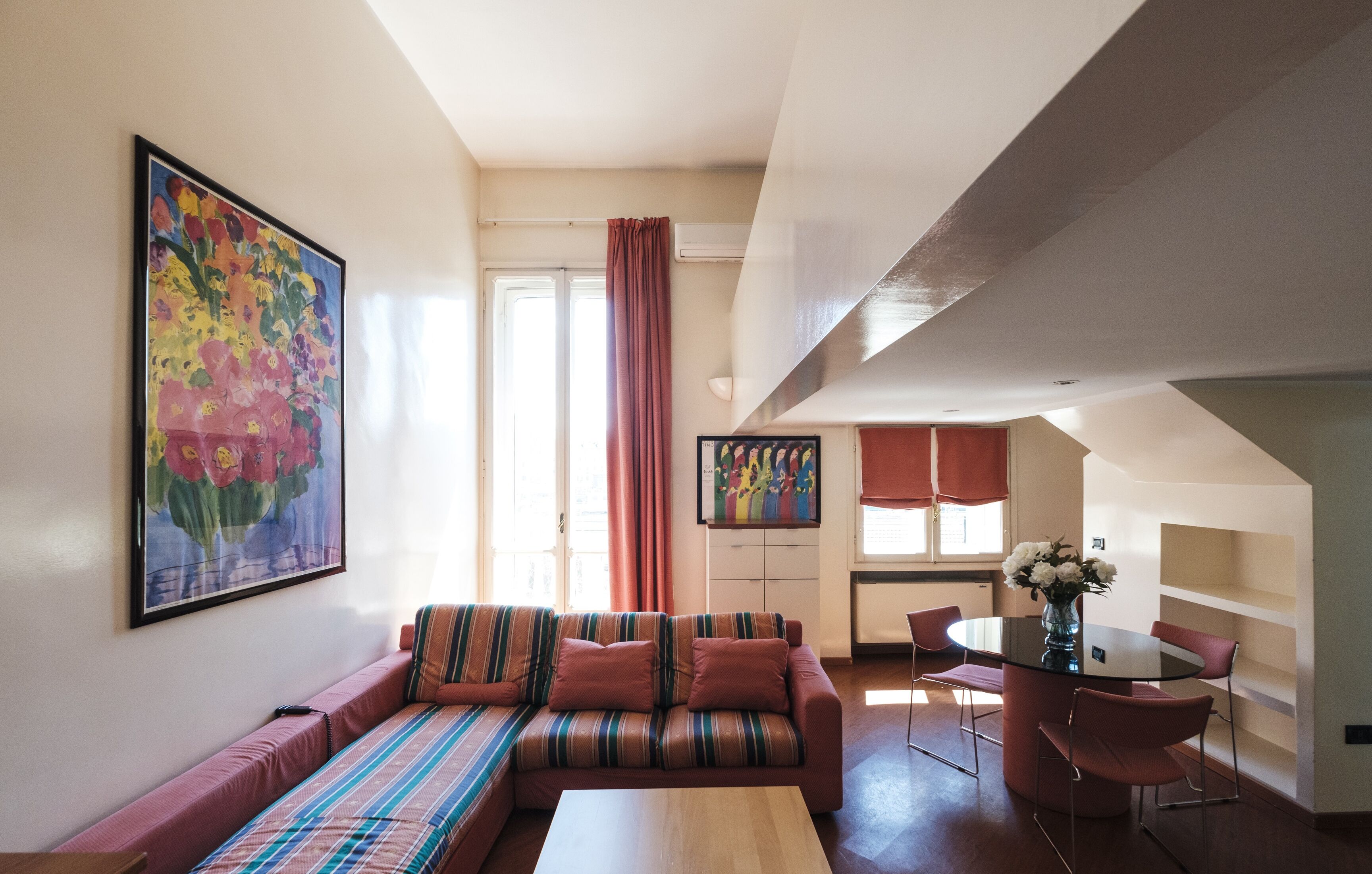 Una sala de estar con un sofá, una mesa de centro y una mesa de comedor con sillas. Hay un cuadro en la pared y una ventana con cortinas.