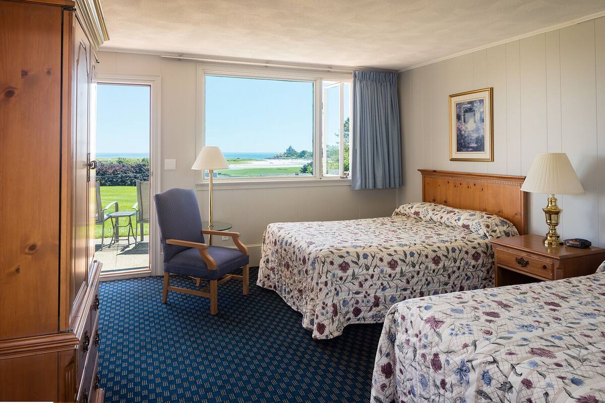 A hotel room with two beds, a chair, a table, and a view of the sea.