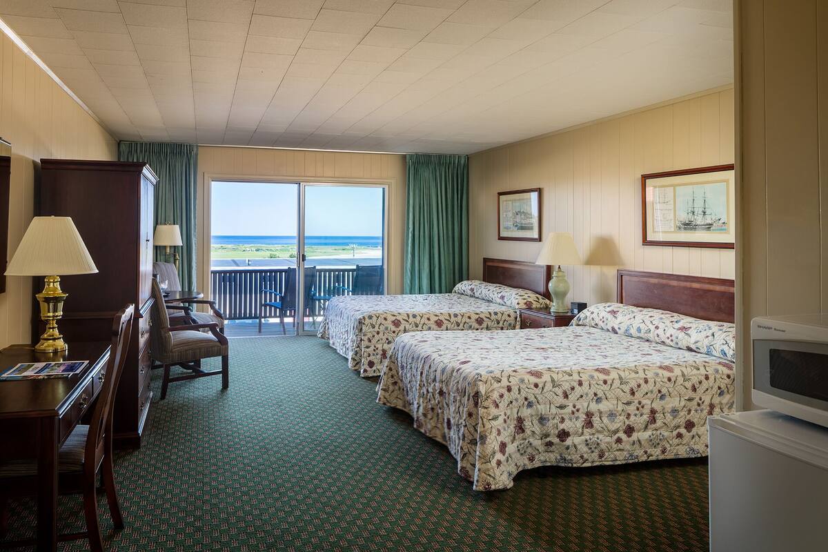 A hotel room with two beds, a desk, a chair, a television, and a balcony with a view of the ocean.