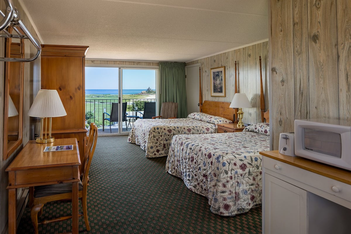 A hotel room with two beds, a desk, a chair, a TV, and a view of the ocean.