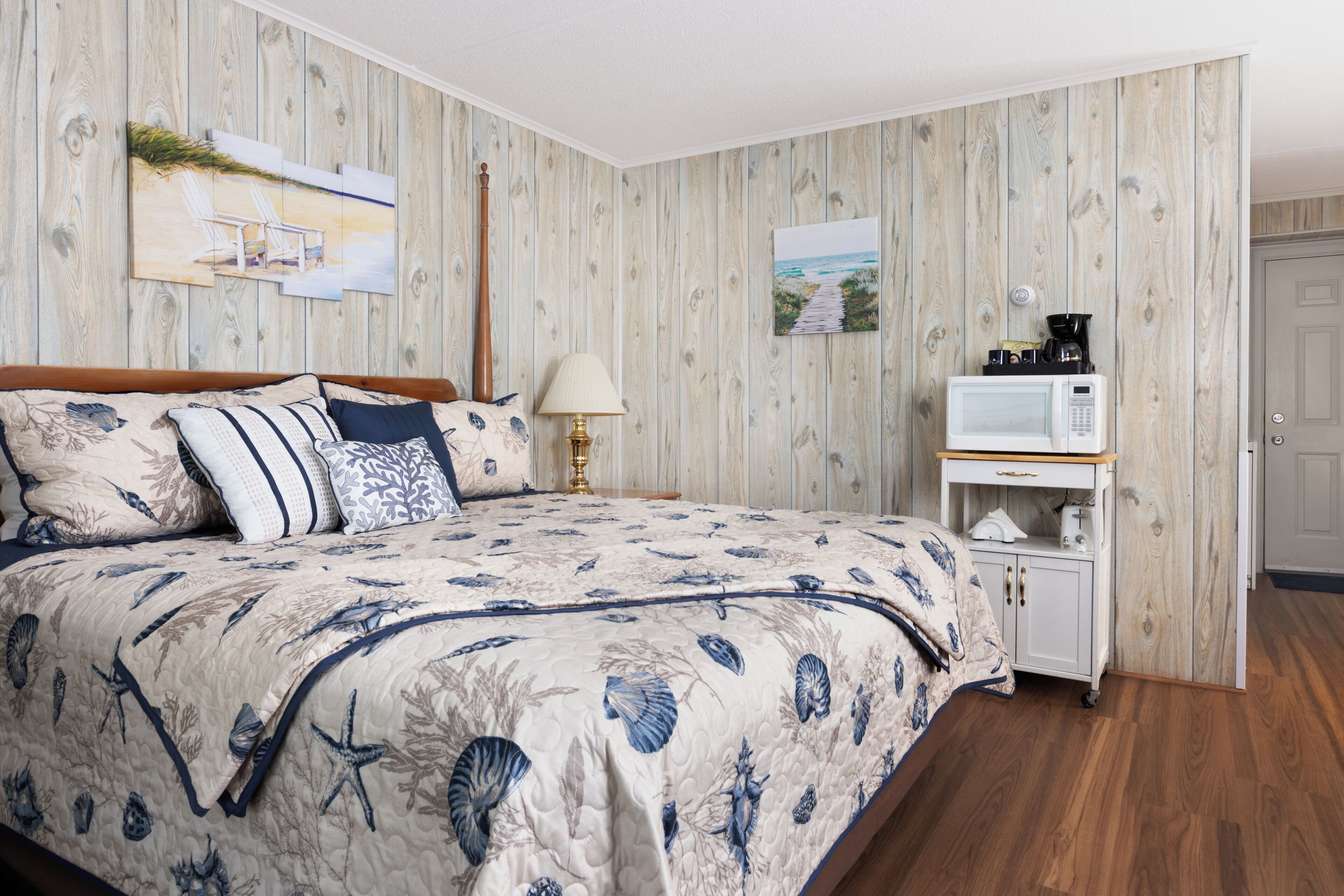 A bedroom with a wooden wall, a bed with a floral duvet, a bedside table with a lamp, and a microwave on a wooden cabinet.