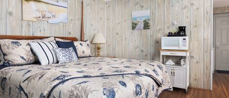 A bedroom with a wooden wall, a bed with a floral duvet, a bedside table with a lamp, and a microwave on a wooden cabinet.