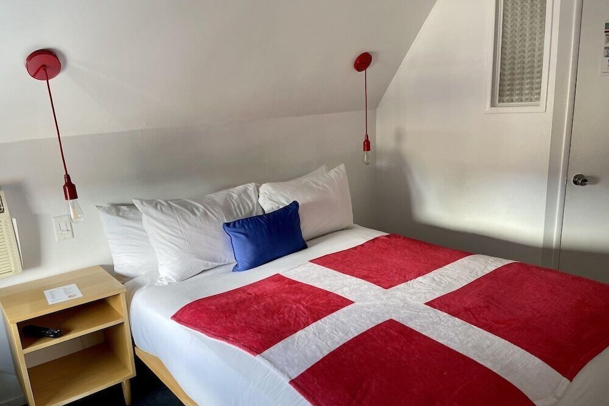 Una cama individual con colcha roja y blanca, una mesita de noche de madera, un almohán azul y una lámpara colgante roja.