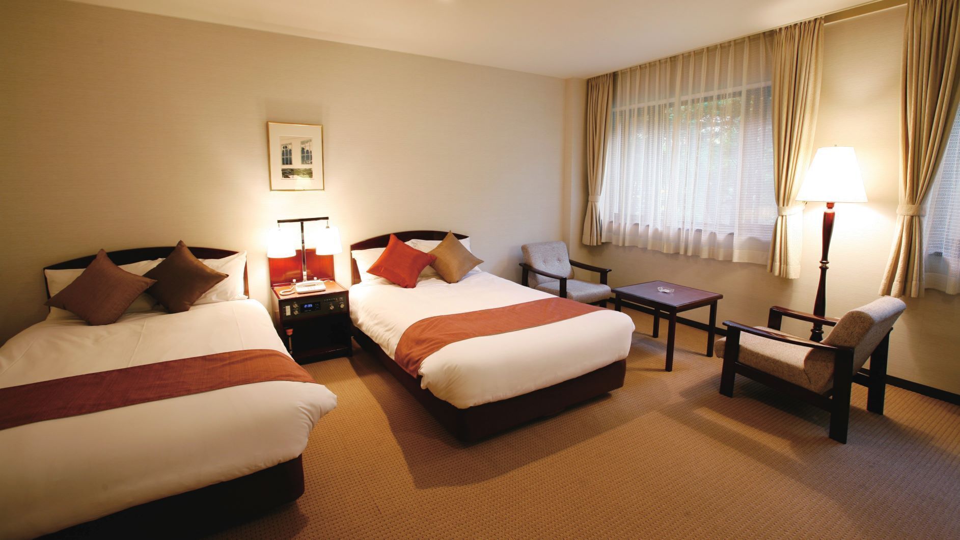 Quarto de hotel com duas camas, uma cadeira, uma mesa pequena e uma janela com cortinas.