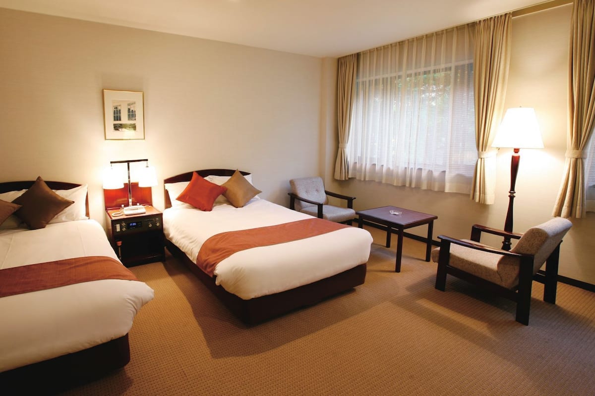 Une chambre d’hôtel avec deux lits, une chaise, une petite table et une fenêtre avec des rideaux.