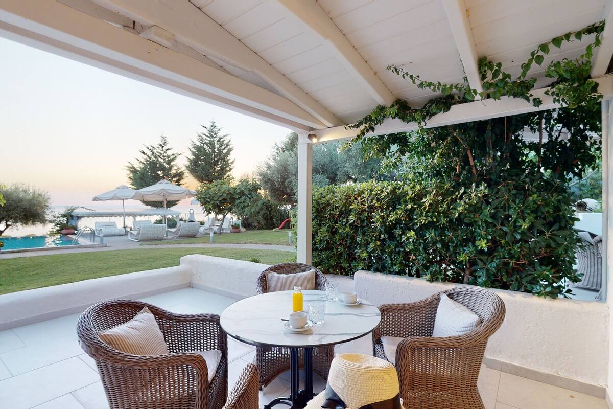 A covered outdoor seating area with wicker furniture, a round table, and a view of a swimming pool and greenery.