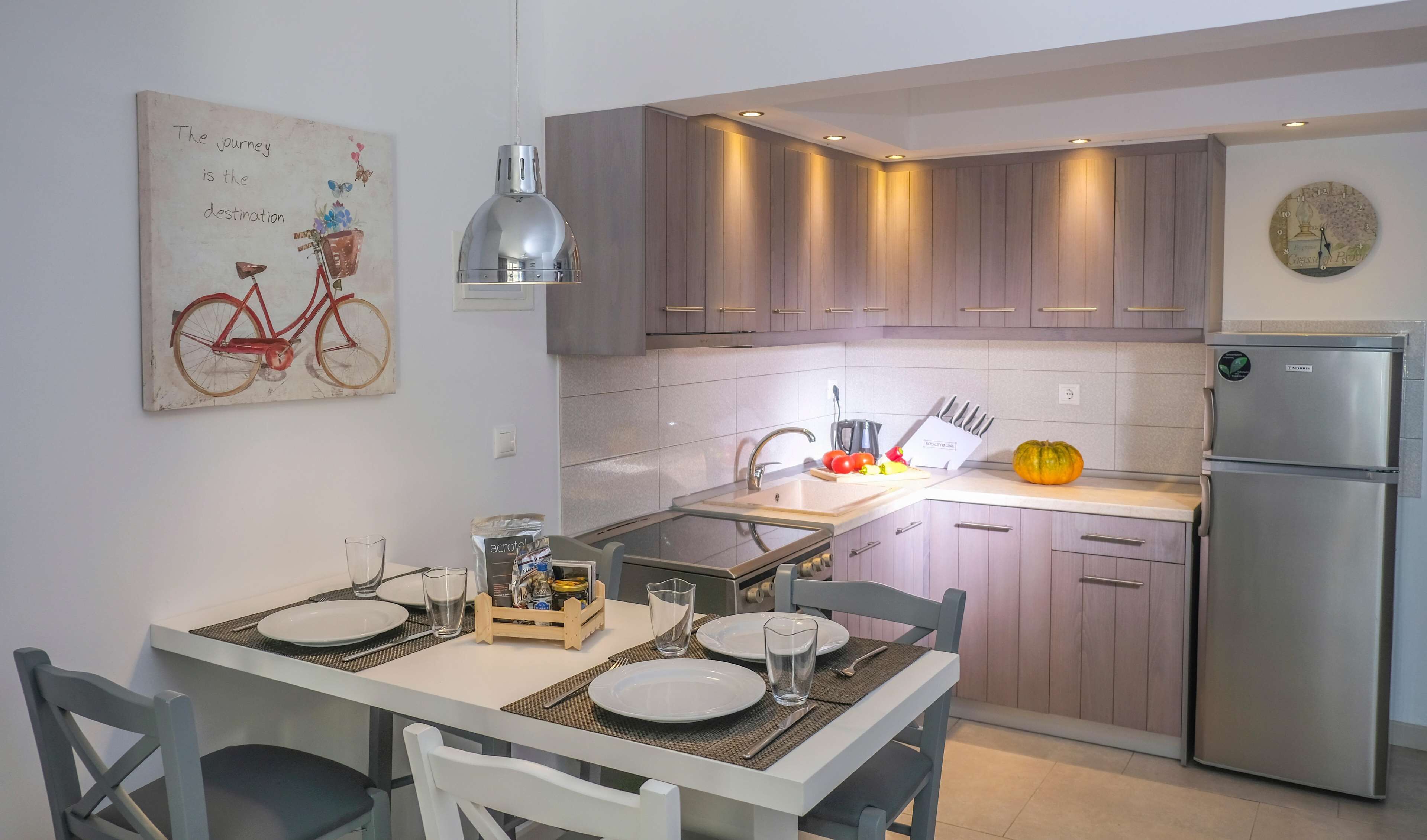 Una cocina moderna con armarios de madera, un refrigerador de acero inoxidable y una zona de comedor dispuesta con platos y vasos.