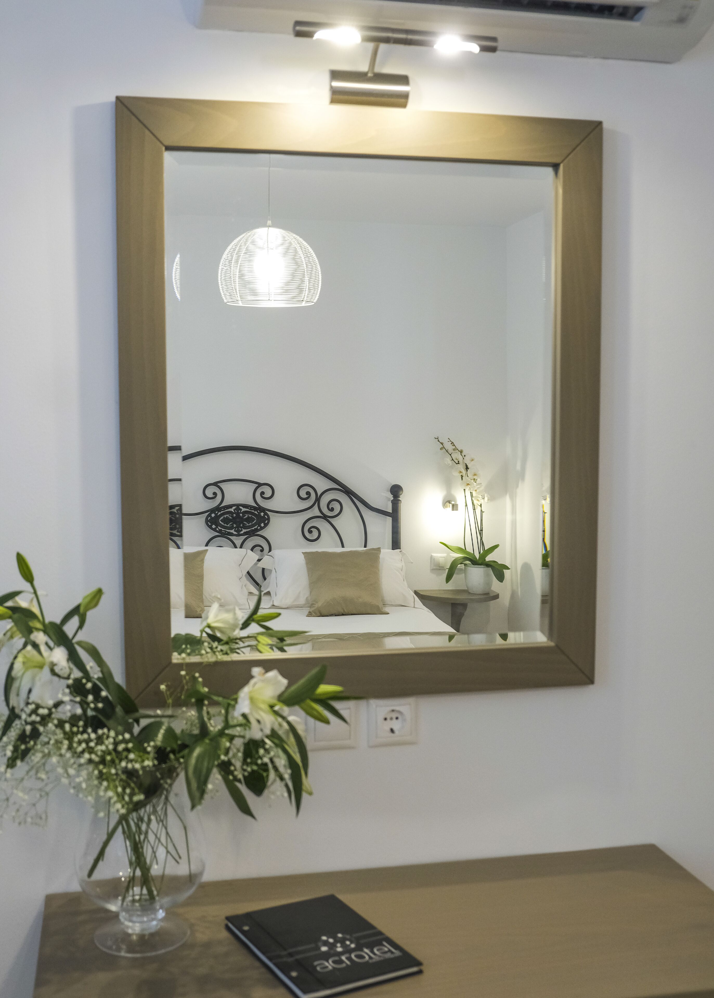 A bedroom with a large mirror, a bed with a headboard, a bedside table with a lamp, and a vase with flowers.