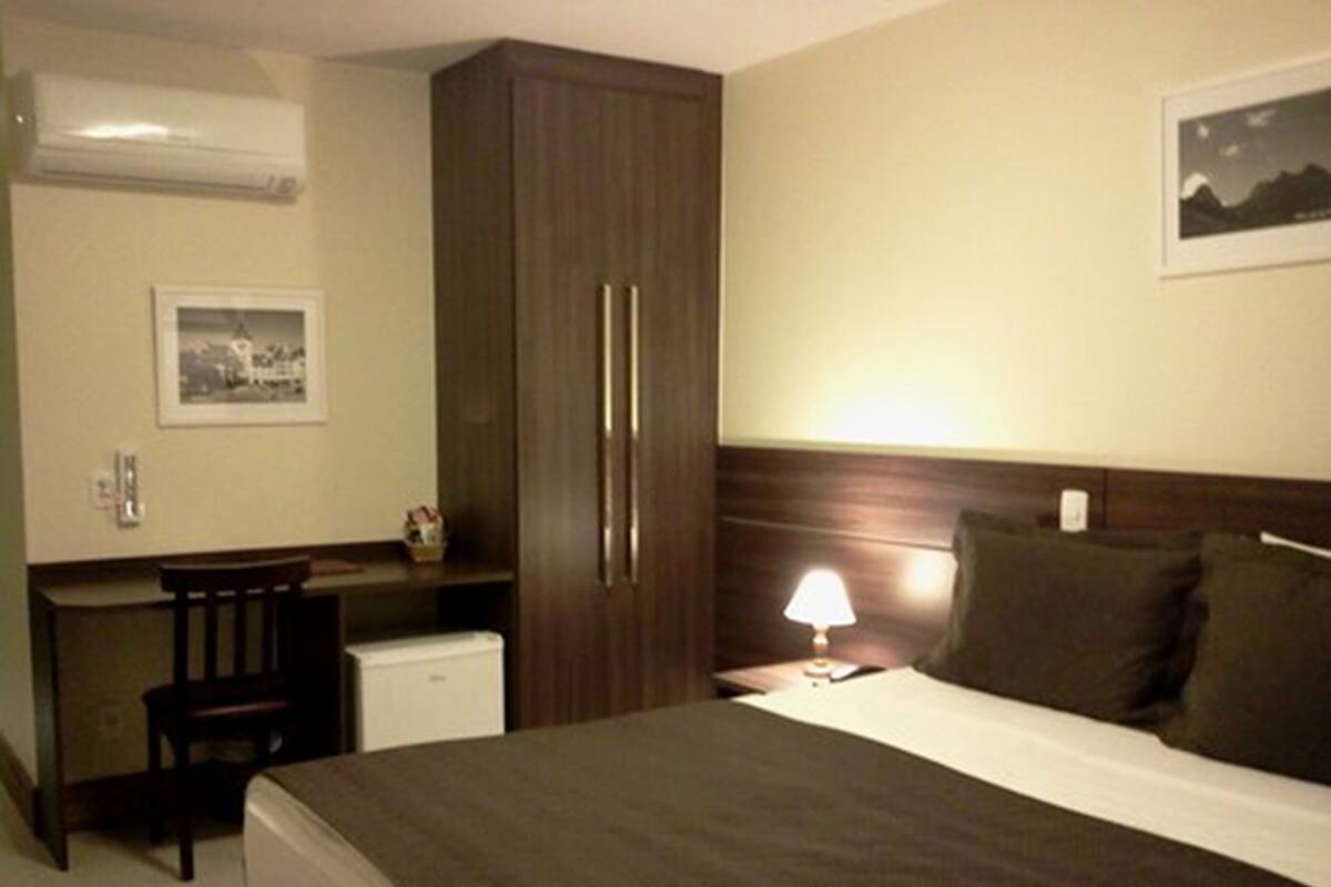 Une chambre d’hôtel comprenant un lit, un bureau, une chaise, une armoire, une table de chevet avec une lampe et un tableau encadré au mur.