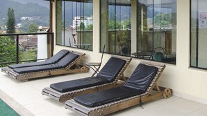 A covered outdoor lounge area with three sun loungers, large windows offering views of the surrounding landscape, and a wooden ceiling structure. - Hotel Willisau (Teresópolis)