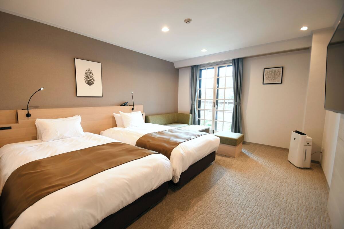 A hotel room with two beds, a desk, a chair, a window with curtains, and a wall-mounted picture.