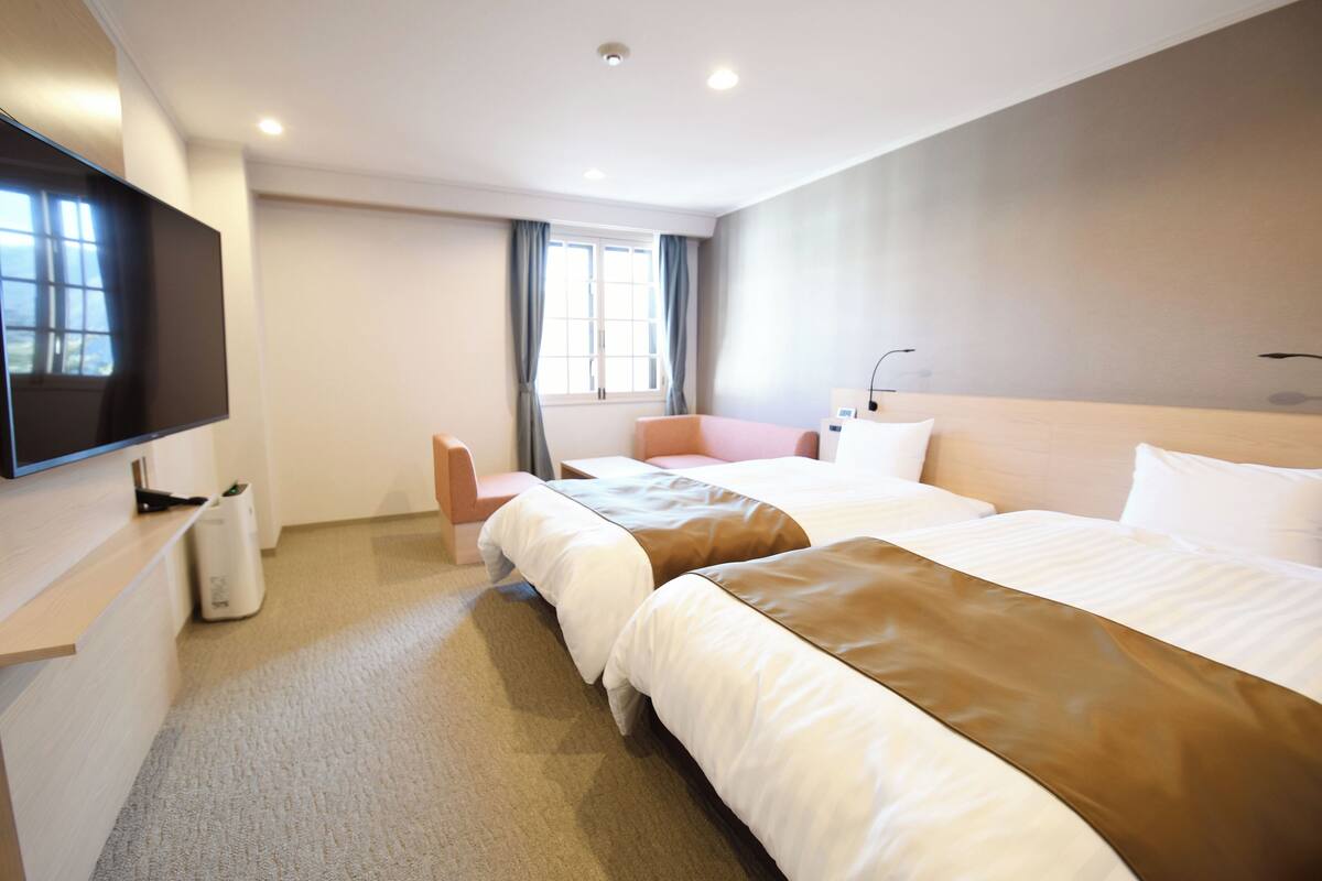 A hotel room with two beds, a TV, a desk, and a window with curtains.
