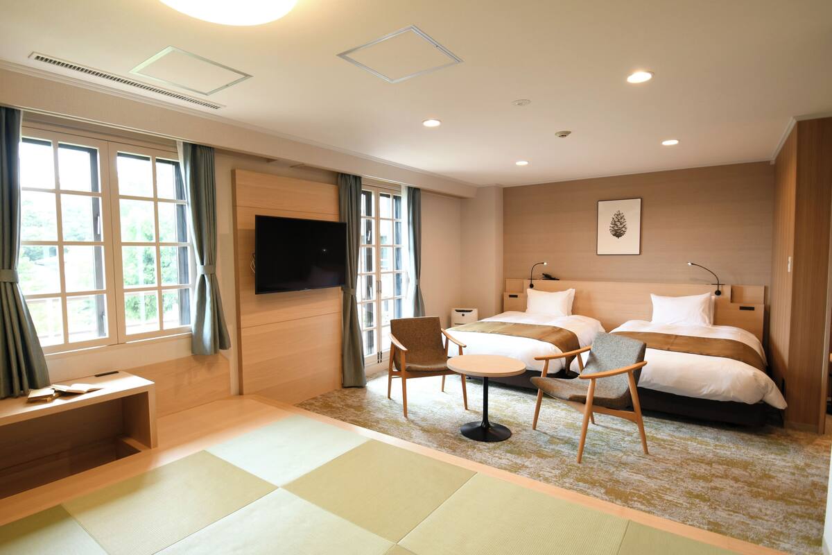 A hotel room with a bed, two chairs, a desk, a TV, and a window with curtains.