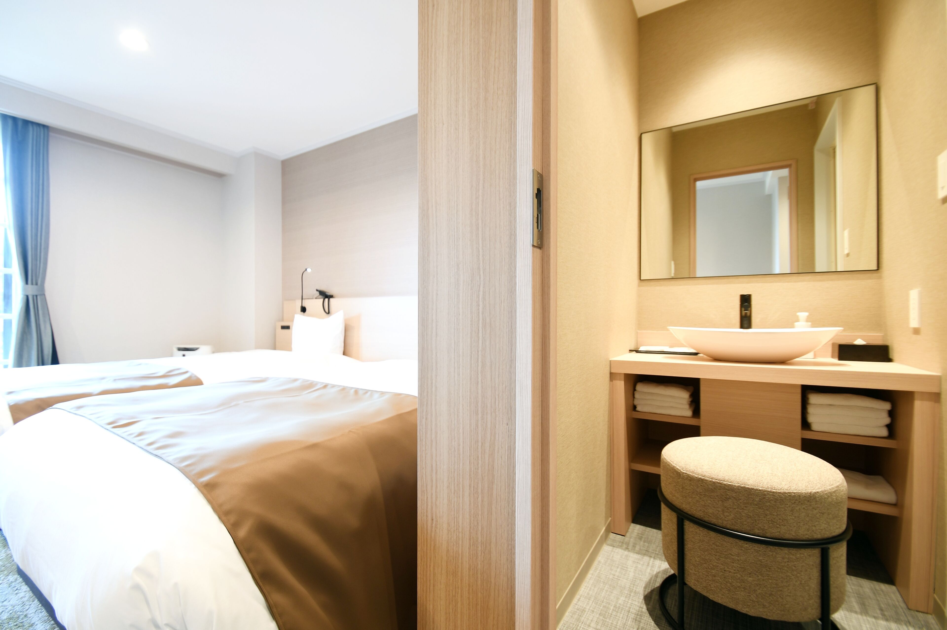 Habitación de hotel con una cama grande, una mesita de noche, un espejo y un baño con lavamanos y espacio para toallas.
