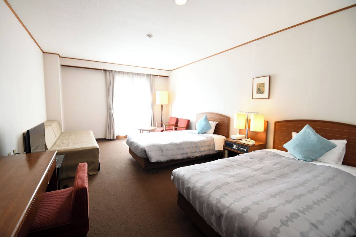 A hotel room with two beds, a desk, a chair, a bedside table with a lamp, and a window with curtains.