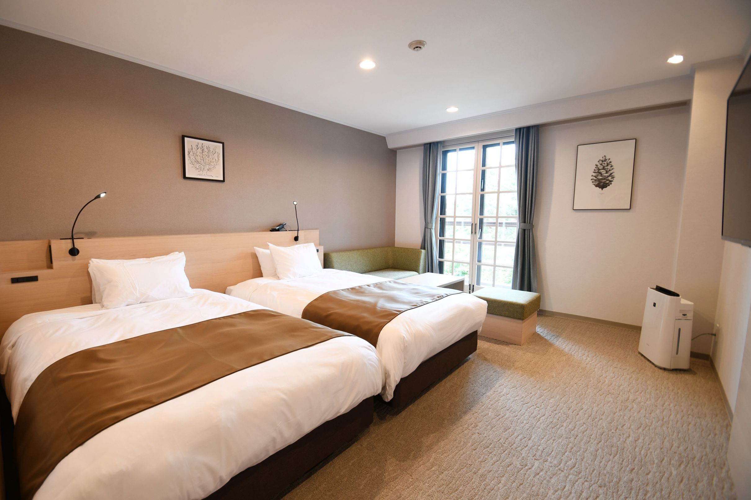 A hotel room with two beds, a desk, a chair, a window with curtains, and a wall-mounted air conditioner.