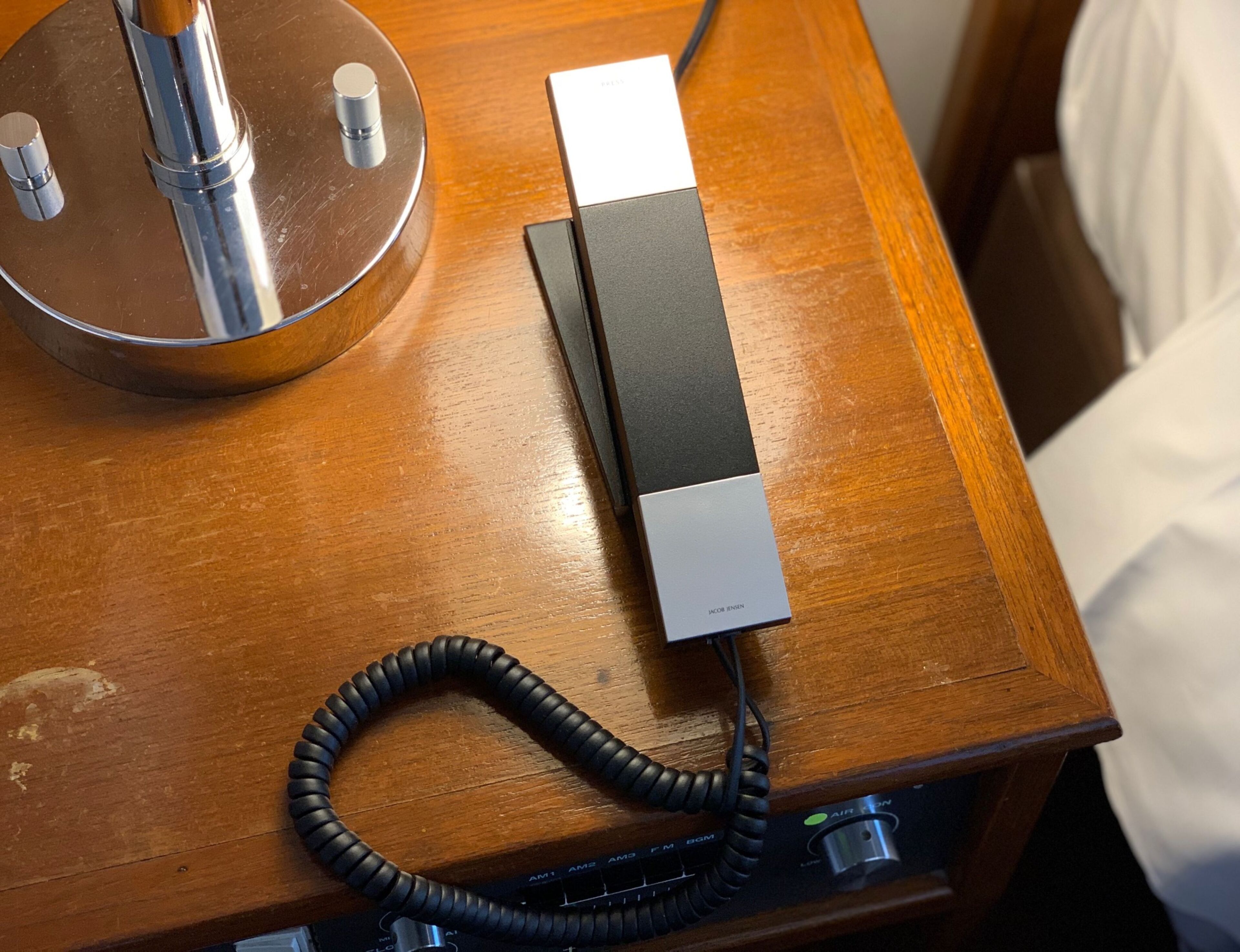 Una mesita de noche de madera con un dispositivo electrónico negro y plateado, posiblemente un cargador, y un cable negro enrollado.