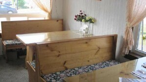 A wooden bench with a floral cushion, a small table with a vase of flowers, and a window with curtains. - Rosecourt Motel (Stratford)