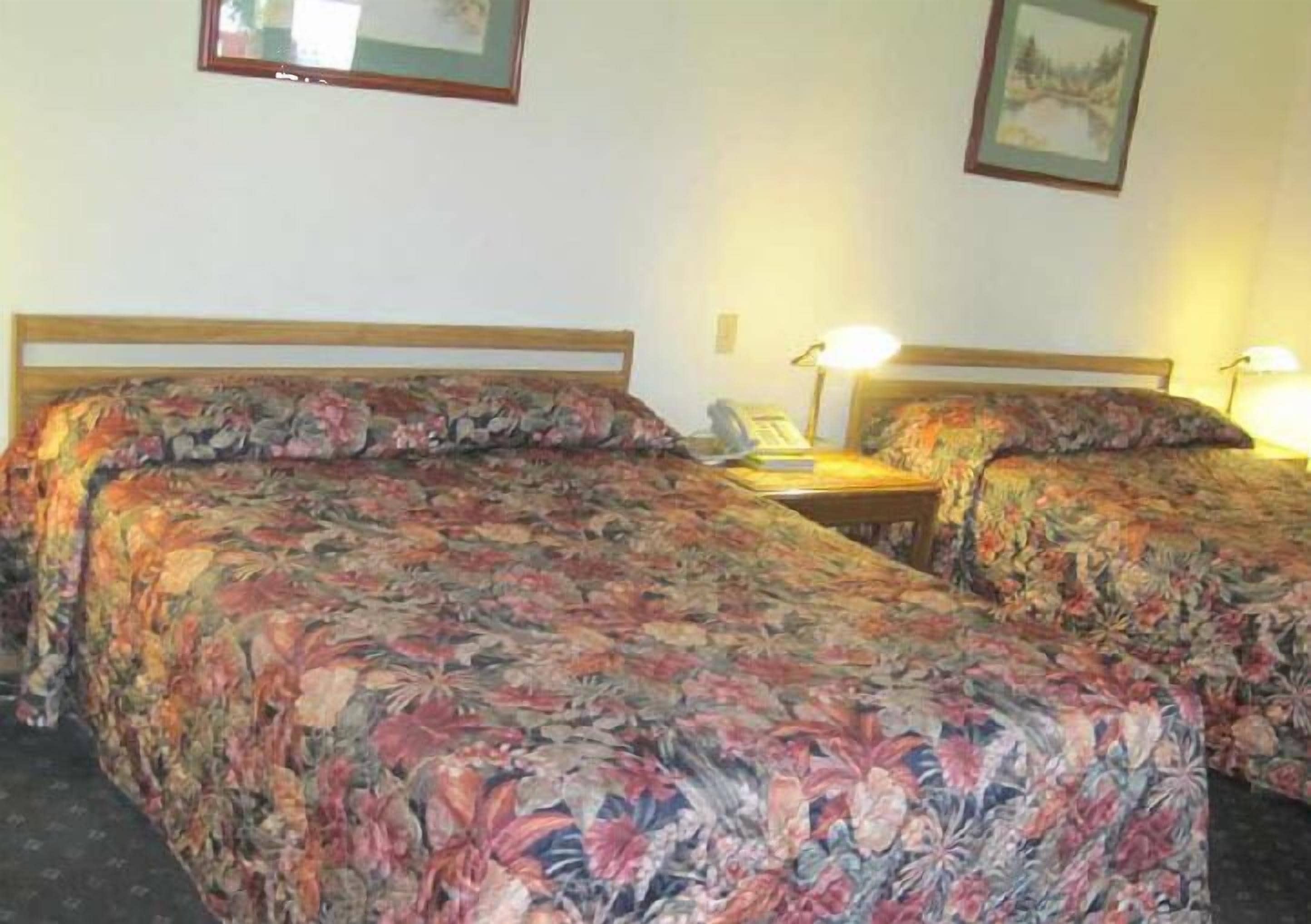 A room with two single beds, each with floral-patterned bedding. There are bedside tables with lamps, and the walls are plain with a framed picture above one bed.