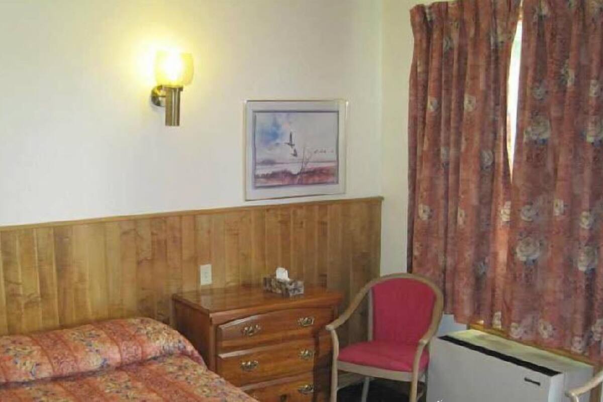 A room with a wooden headboard, a bed with a floral bedspread, a wooden dresser, a red chair, a desk with a lamp, and a window with curtains.