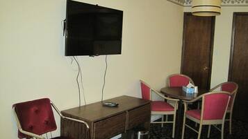 A room with a TV mounted on the wall, a wooden desk with a remote control, and two red chairs around a small table.